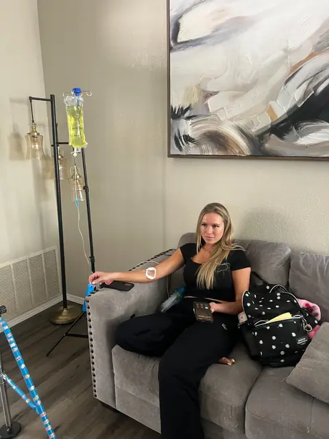 A client sitting on a gray sofa at home receiving an IV drip from a portable IV stand