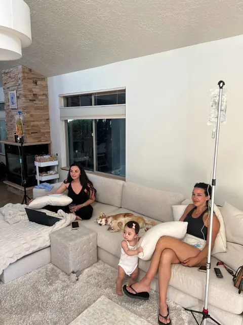 Two women receiving IV drips on a sectional sofa at home with a toddler beside them