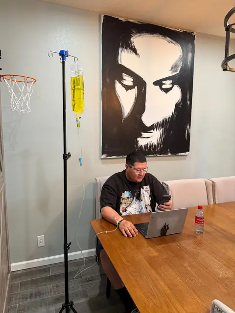 A man working on a laptop at his dining table while receiving an IV drip at home